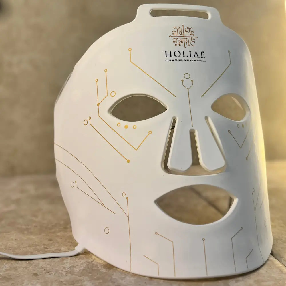 Masque LED visage Holiaē RĒ:GEN I, vue 3/4 sur fond minéral, circuits dorés.